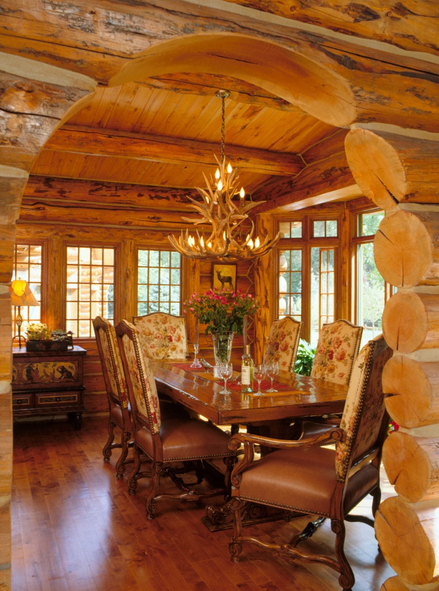 A rustic dining room in a log cabin. The room is constructed of large, exposed logs. The ceiling is made of wood planks, and there's an arched doorway framing the scene. A large, dark wood dining table is centrally located, surrounded by ornate chairs with floral upholstery. 