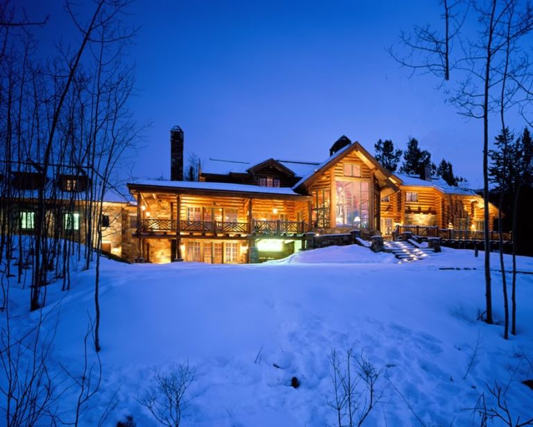 luxurious log cabin nestled in a snowy landscape at twilight. The log cabin features large windows, especially a prominent wall of windows on the back, showing interior lighting.